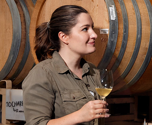 amanda gorter - woman with a dark brown ponytail and a olive green button down shirt smiling in front of wine barrels holding a glass of wine