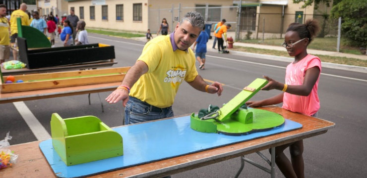 man instructs young girl how to play a frog launch game on a table out in the street