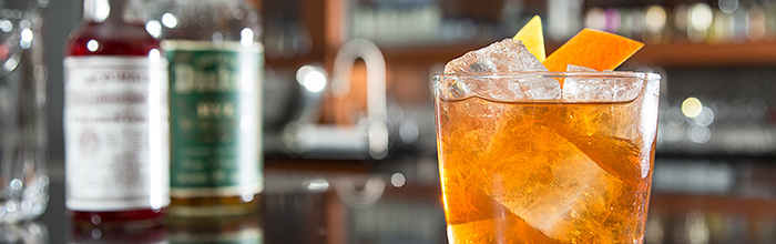 orange-hue cocktail with large ice chunks garnished with an orange peel overshadowing glass liquor bottles in the background