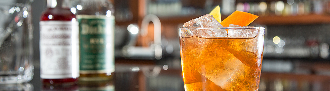 orange-hue cocktail with large ice chunks garnished with an orange peel overshadowing glass liquor bottles in the background