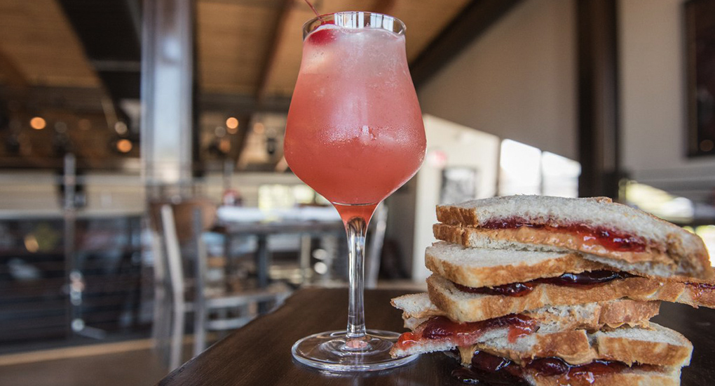 Pinkish colored Dogfish Head cocktail in wine glass adjacent to stacked peanut butter jelly sandwiches