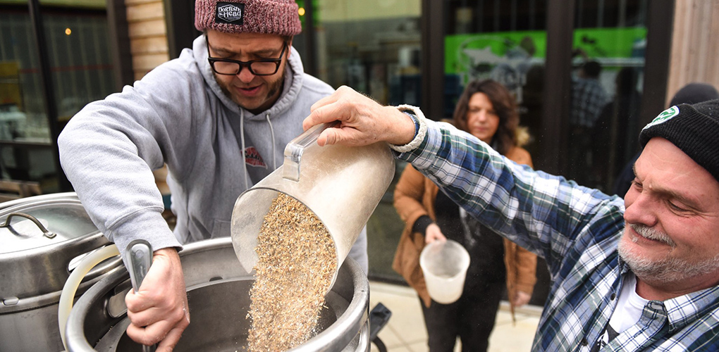 Two male Dogfish Head employees pouring hops into a keg as a woman looks on from the background