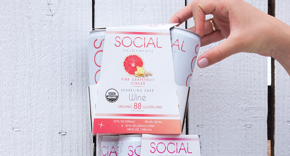 a hand pulling a white can of SOCIAL wine out of packaging in front of a wood fence