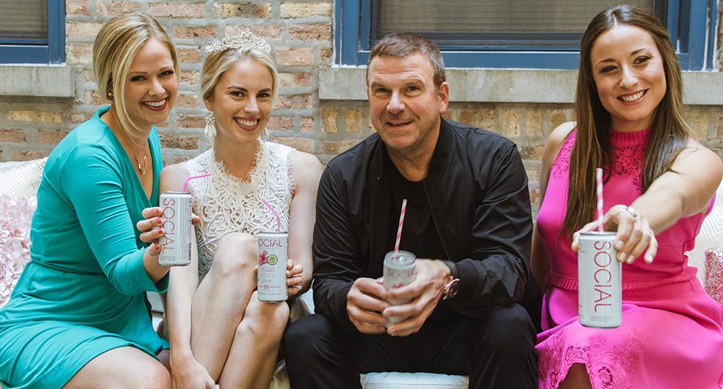 3 happy women and a smiling man sitting on a couch holding white cans of SOCIAL wine with straws in them