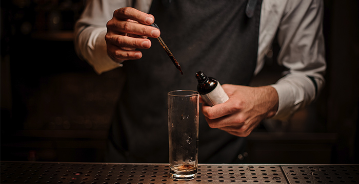 mixologist dripping bitters into an empty highball cocktail glass on a steel counter