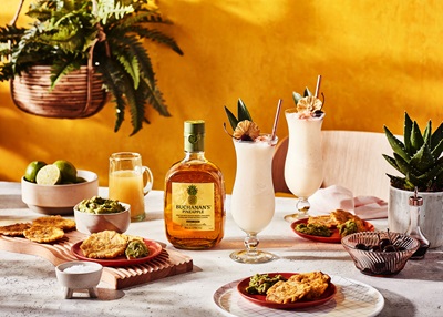Bottle of Buchanan's Pineapple Scotch surrounded by food and cocktails against a yellow wall with a plant hanging in the left corner