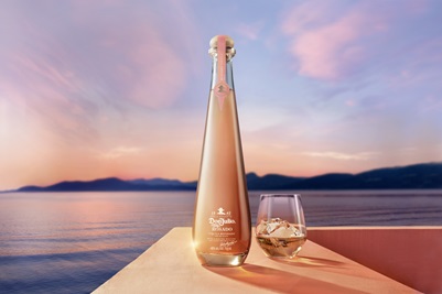 Don Julio Rosado in center of screen sitting atop a ledge with the sunset over water in the background. A glass is next to the bottle. 