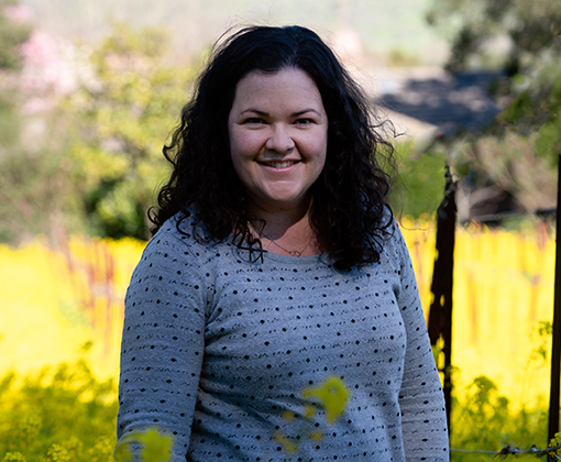 anne dempsey - smiling woman with dark curly shoulder length hair wearing a light blue shirt with dark blue polka dots standing in a green-leafed vineyard