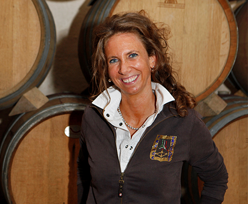 barbara tamburini - woman with dark blonde curly long hair wearing a white button down top and a brown sweater standing in front wine barrels