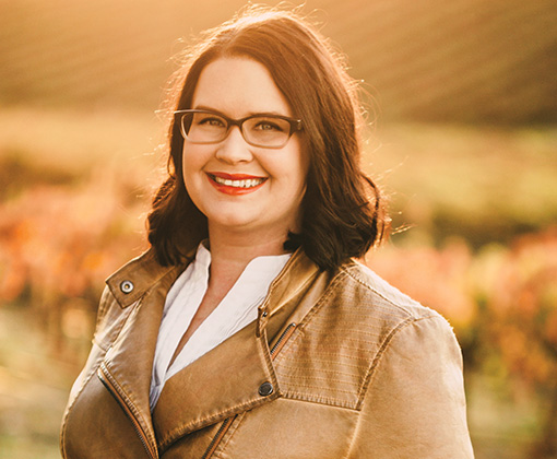 emily haines - woman with shoulder length brown hair wearing glasses and a leather jacket with a white shirt smiling standing in a field looking in an eastern exposure