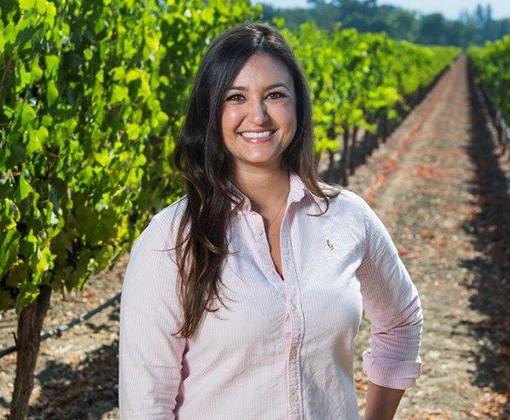 brunette woman in a rose-colored blouse standing in a green-leafed vineyard