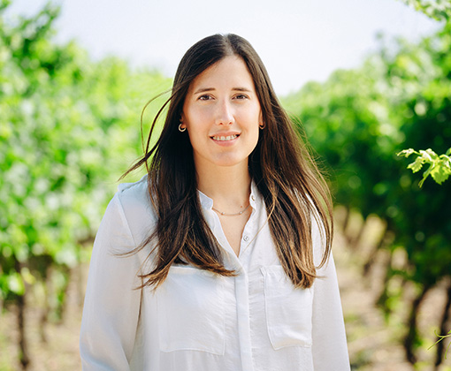 magdalena viani - woman with long brown hair and a white button down shirt standing in a green-leafed vineyard looking at the viewer