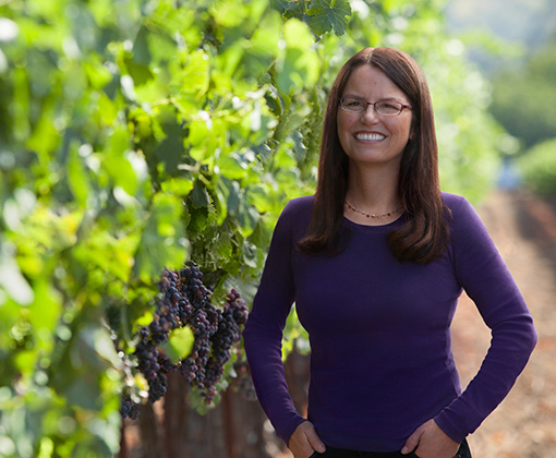woman with glasses wearing a violet shirt in a green-leafed vineyard