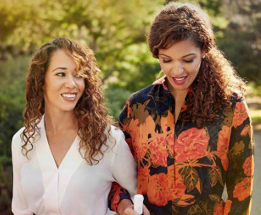 A woman with long curly light brown hair and a white button down top standing arm-in-arm with a taller woman with curly dark brown hair pulled in a ponytail wearing a floral pattern top in a green-leafed vineyard